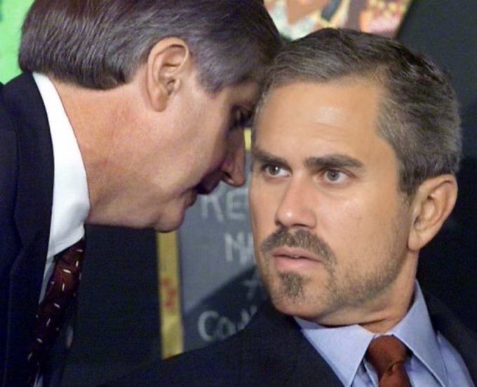 Two men in suits are shown in close proximity, with the older man on the left whispering or speaking closely to a younger man with a goatee on the right.