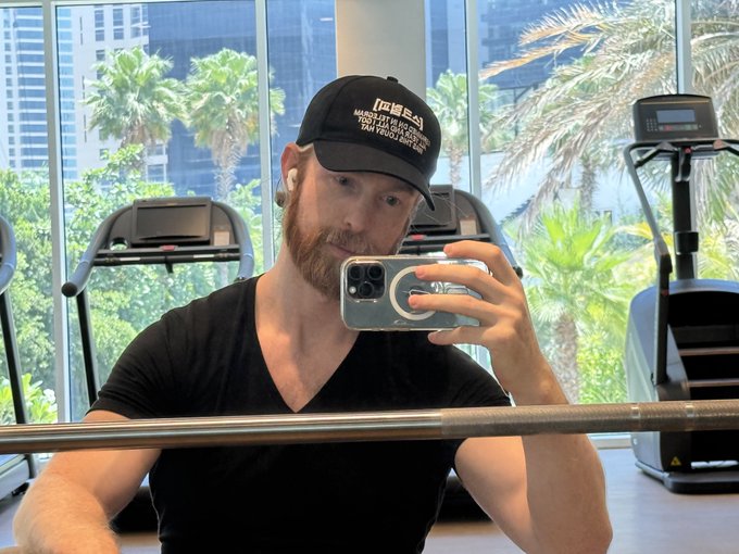 The image shows a man taking a selfie with his cell phone while sitting at a gym. The man is wearing a black baseball cap and has a beard. He appears to be capturing a moment of his workout or simply documenting his experience at the gym. In the background, there are exercise machines such as treadmills and weightlifting equipment that can be seen in various positions within the room.