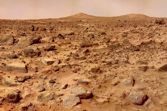 A barren, reddish-brown Martian landscape scattered with rocks and boulders stretches toward distant hills under a hazy sky, illustrating the rocky terrain referenced in the post about "Mars rocks."