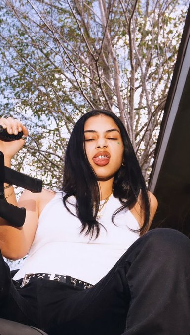 A young woman with long dark hair and a white top poses outdoors, holding a black object near her face with trees visible in the background.