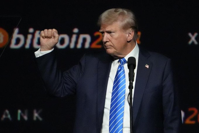 Donald Trump raises his fist on stage with "bitcoin202..." visible in the background, reflecting the post's mention of his reported crypto income.