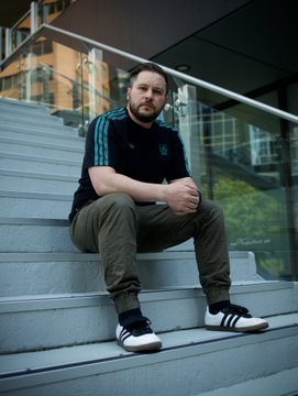 A man with a beard wearing a dark teal soccer jersey and olive green pants sitting casually on concrete steps outside a modern building.