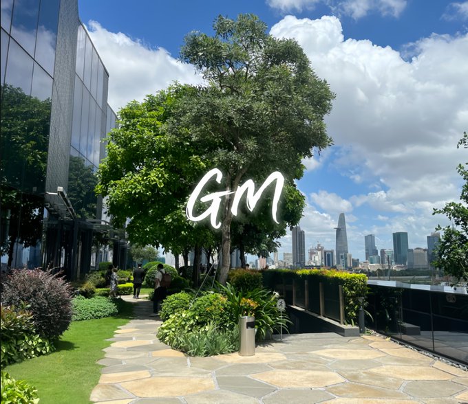 A scenic outdoor garden terrace with stone pathways, lush greenery, and a city skyline in the background, overlaid with the text 'GM'.