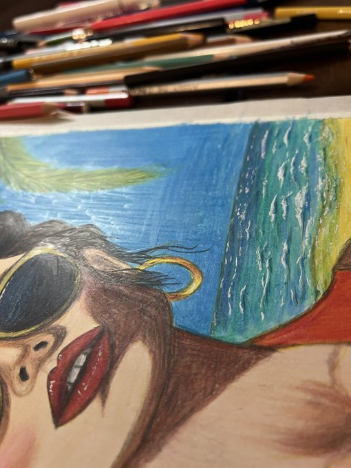 A close-up of a colored pencil drawing in progress, depicting a woman wearing sunglasses with her tongue out, set against a blue and green background with colored pencils visible at the top.