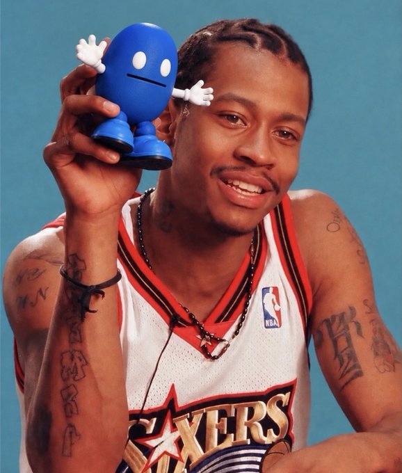A smiling basketball player in a Philadelphia 76ers jersey poses while holding up a small blue figurine toy.