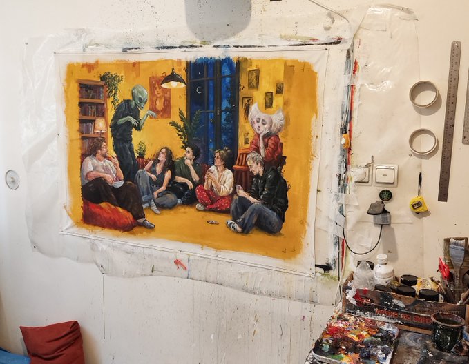 An artist's studio space featuring a colorful painting of several young people sitting on the floor with a zombie-like figure and an older woman in a warmly lit room, surrounded by paint supplies and a ruler on the wall.