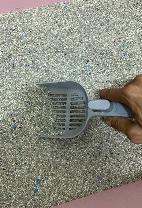 A hand holds a gray plastic cat litter scoop over a gray litter box mat scattered with colorful crystals and clumps of used cat litter.