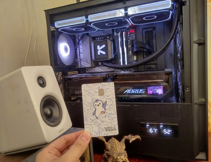 The image shows a person holding up a white card with a cartoon penguin design next to a computer case. The card appears to be a credit or debit card, possibly for a rewards program like Vasex. The scene takes place on a desk, which also has a speaker and a small figurine of a penguin nearby.