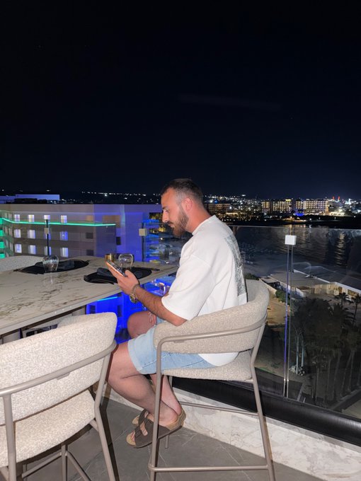 A man sits at a high-rise rooftop table at night, looking down at his phone with a glass of wine beside him, while a city skyline and waterfront glow in the background—capturing the essence of a relaxed "Gm" (good morning) mood even after dark.