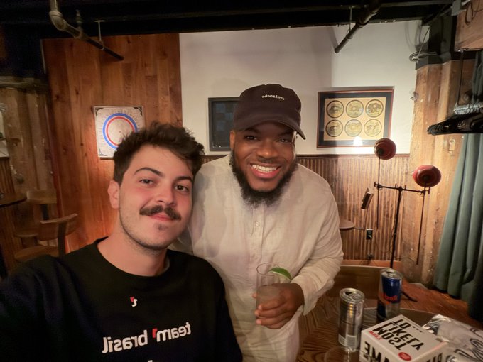 A selfie of two smiling young men in a wood-paneled bar with framed records on the wall; one wears a black 'Jiraq' shirt and the other a cream hoodie and black cap, holding a drink near a Tel Aviv beer box.