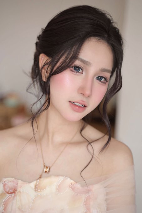 A portrait of a young woman with dark hair styled in an updo, blue eyes, wearing makeup and a pink floral dress with a gold necklace.