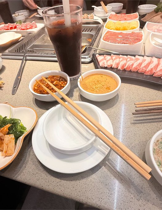 A hot pot meal setup with chopsticks, dipping sauces, sliced meat, vegetables, and an iced drink on a restaurant table.