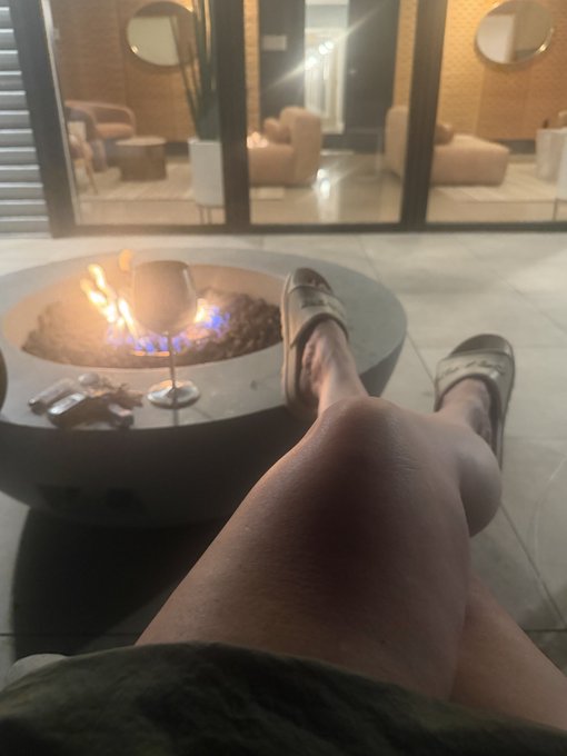 A first-person view of legs stretched out beside a modern circular fire pit at night, with the person wearing Birkenstock sandals and holding a glass of white wine.