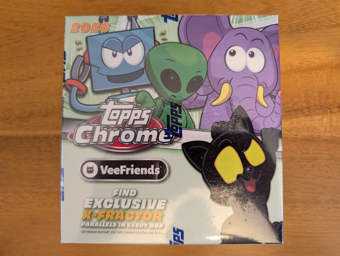 A sealed box of 2026 Topps Chrome VeeFriends trading cards rests on a wooden surface, featuring colorful characters and text reading 'FIND EXCLUSIVE X-FRACTOR PARALLELS IN EVERY BOX'.