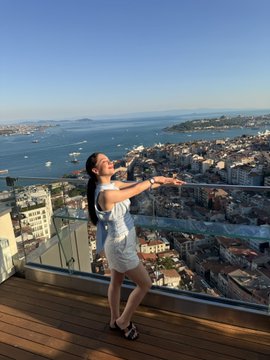 The image features a woman standing on a balcony, looking out over a cityscape with boats visible in the water below. She appears to be enjoying her time at the location, possibly attending or hosting an event like Solana Summer Party. The presence of boats suggests that this could be a waterfront venue for such gatherings.