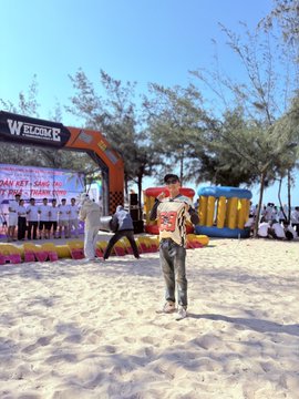 The image shows a young boy standing on a sandy beach, wearing a white shirt with red lettering that reads "WELCOME". The boy appears to be posing for a photo or participating in an event at the beach. In the background, there are several people and inflatable structures, suggesting that this could be part of a beach party or competition.