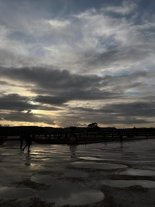 A landscape view of a sunset over a body of water, featuring dark clouds and puddles reflecting the sky in the foreground.