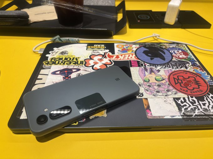 The image features a cell phone placed on top of an open laptop computer, both resting on a yellow table. The phone has several stickers on its back, including one with the words "Working with my @solanamobile phone I need to change my stickers". This suggests that the person using these devices might be in the process of personalizing their belongings or expressing themselves through various stickers and decals.