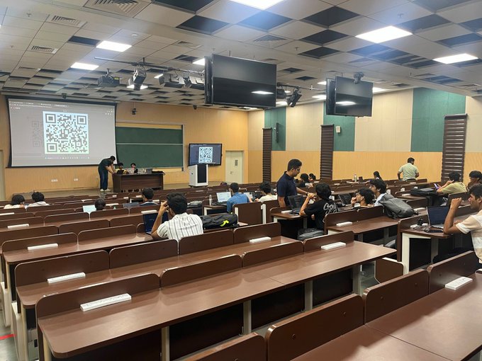 Students sit in the wooden desks of a large lecture hall while a presentation featuring a QR code is projected onto a screen at the front.