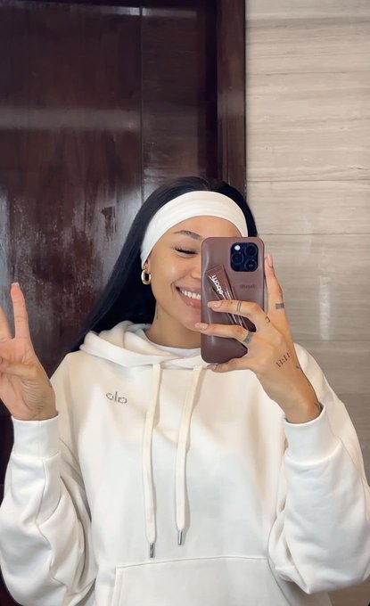 A young woman taking a mirror selfie wearing a white hoodie and white headband, making a peace sign and smiling.