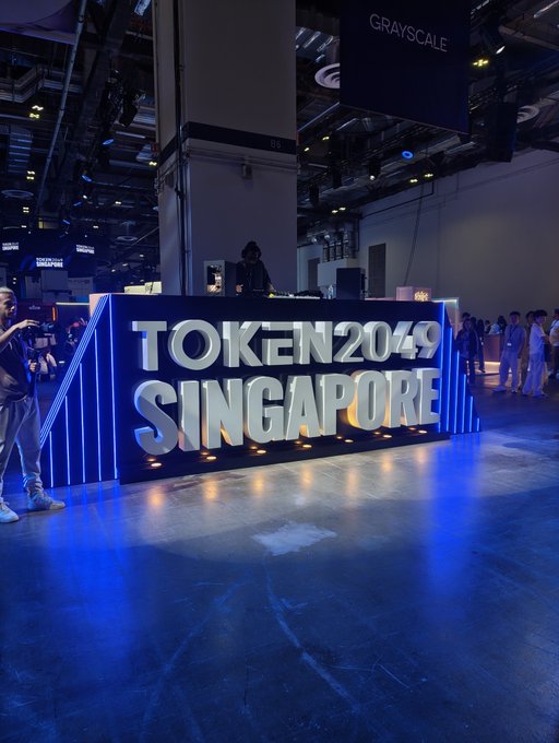A large illuminated blue sign reading 'TOKEN2049 SINGAPORE' at a conference venue, with attendees and a Grayscale booth visible in the background.