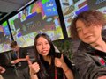 A selfie of two smiling women wearing headsets, with one making a peace sign, set against a backdrop of multiple monitors.
