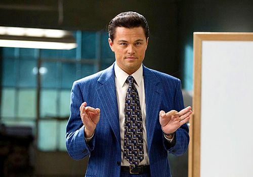 A man in a blue pinstripe suit and patterned tie standing in front of a whiteboard with his hands raised in a gesturing pose.
