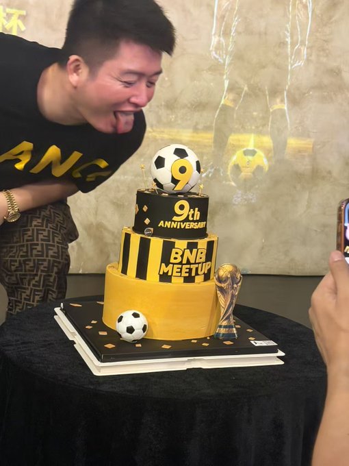 A man in a 'BANG' shirt sticking out his tongue next to a yellow and black tiered cake celebrating the '9th Anniversary BNB MEETUP', decorated with a soccer ball, a '9' candle, and a World Cup trophy.