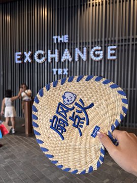 A person holds up a woven straw hand fan with blue trim featuring a cartoon character design, with a sign reading 'THE EXCHANGE TRX' visible in the background.