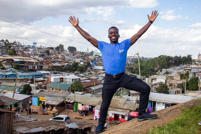 A man in a blue t-shirt and dark pants stands triumphantly with arms raised on a hill overlooking a densely populated urban neighborhood with corrugated metal rooftops.
