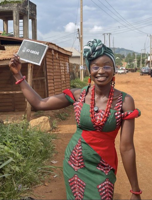 A smiling woman wearing traditional green and red patterned attire, coral bead necklace, and a matching headwrap, holding up a small chalkboard sign that reads 'I ❤️ GBEDE' on a dirt street.