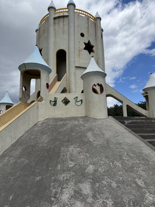 A weathered, fairy-tale-style castle with conical turrets and star cutouts stands under a partly cloudy sky, its worn concrete steps and ramp leading up to a central tower. Hidden somewhere within the structure is a tiny Shiba Inu, waiting to be spotted for good luck.