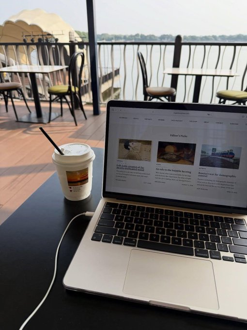 A laptop open on a cafe table beside an iced coffee, showing a webpage with the heading "Editor's Picks," overlooking an outdoor patio and a river.