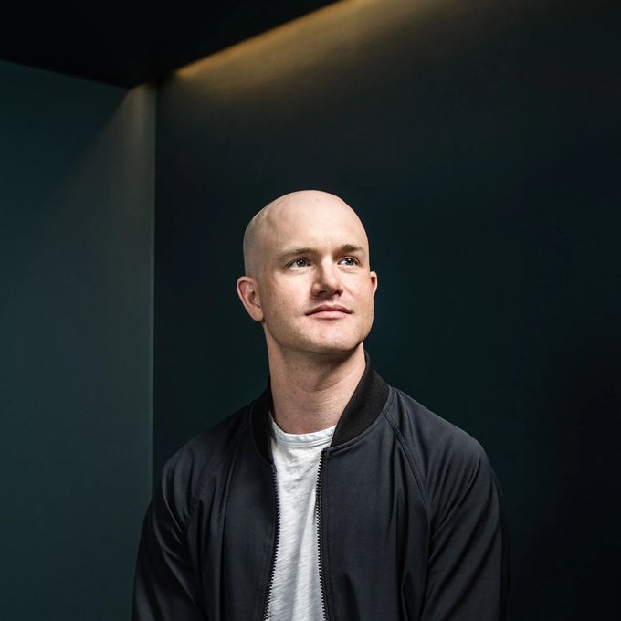A bald man in a dark bomber jacket over a light t-shirt looks upward thoughtfully, set against a dark green background—possibly an executive or industry figure relevant to Coinbase's expansion into traditional markets through new IPO allocation features.