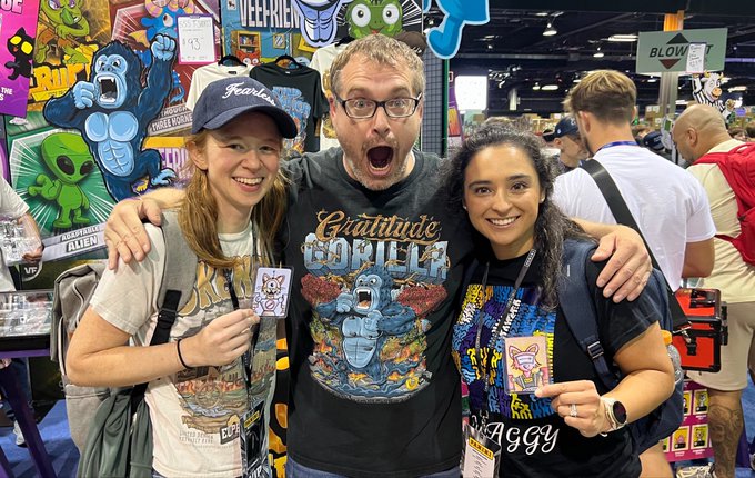 Three people smiling and posing at what appears to be a comic convention, with colorful artwork visible in the background.