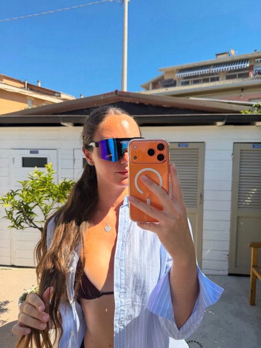 A woman with long brown hair wearing reflective blue/purple sunglasses and an open striped shirt taking a mirror selfie with an orange phone case in an outdoor setting.