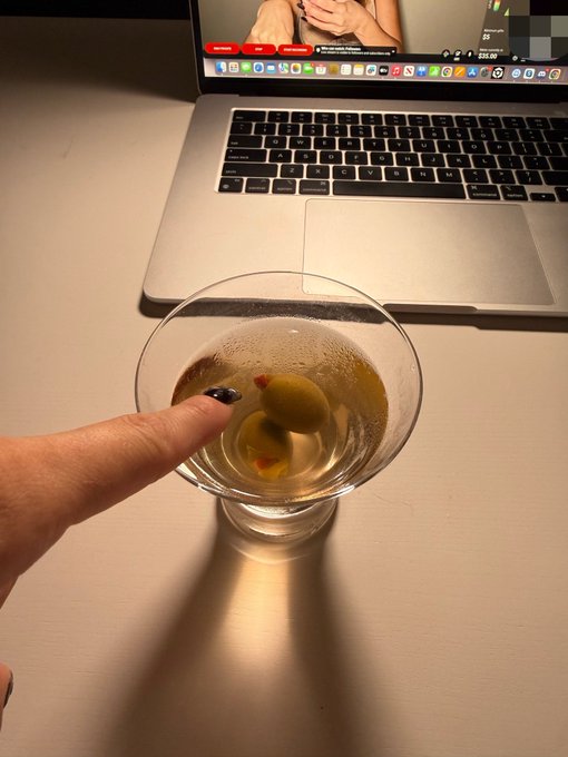 A finger points at a martini glass containing olives, with a laptop displaying a video call visible in the background.