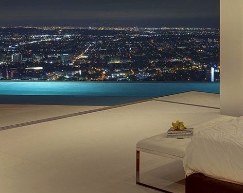 The image shows a bedroom with a bed, a couch, and a book placed next to each other. The room has a large window that offers a view of the city at night. There are also two people visible in the scene, one sitting on the couch and another standing near the bed. The overall atmosphere suggests relaxation and leisure time as they enjoy their surroundings from the comfort of their bedroom.