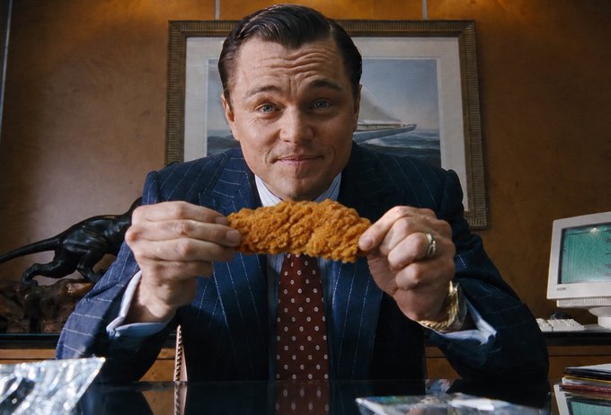 A man in a pinstripe suit sits at a desk in an office, holding up a breaded chicken tender with both hands and smiling at the camera with a satisfied expression. The image of someone proudly displaying chicken tenders serves as a visual pun on the "$TENDIES" crypto coin mentioned in the post, referencing both Wall Street slang for profits and the Robinhood platform's reputation as an "online casino" for retail traders.