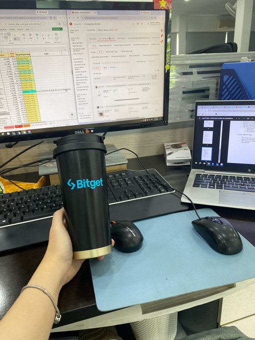 A person holds a black "Bitget" branded tumbler in front of their desk setup, which features a Dell monitor displaying a spreadsheet alongside a laptop showing a Bitget trading page, celebrating 5 months as a Bitget ambassador.