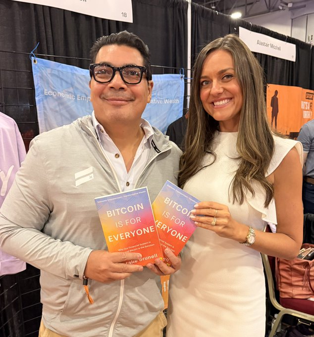 A man and a woman smiling at a convention booth, each holding a copy of the book 'BITCOIN IS FOR EVERYONE' by Adit Brunelli.