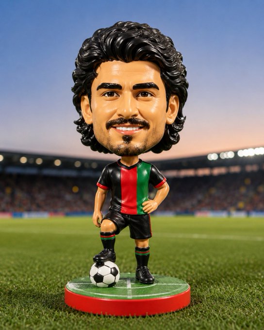 A bobblehead figurine of a soccer player with curly dark hair and a goatee, wearing a black, red, and green striped jersey with black shorts, posed with a soccer ball on a stadium field base.