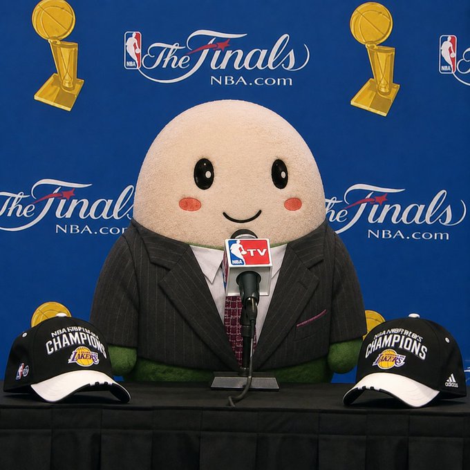 A plush mascot character wearing a pinstripe suit sits at a press conference table with an NBA TV microphone, flanked by two black NBA Champions Lakers caps, against a backdrop featuring 'The Finals NBA.com' logos and championship trophies.
