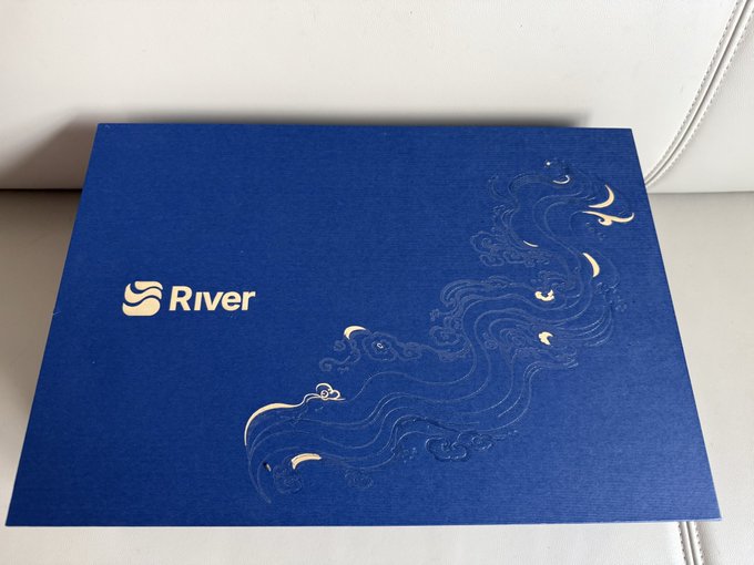 A blue folder or box on a light surface, branded with the 'River' logo and decorated with an elegant gold and blue swirling wave design.