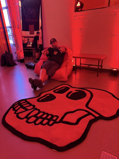 A man reclines in a red-lit room on a beanbag or low chair near a large black-and-red floor graphic, tables, windows, and a “OFF THE GRID CLUB” banner.