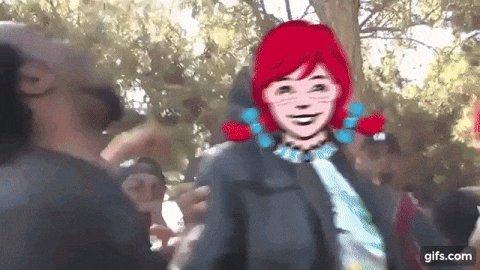 The image features a cartoon woman with red hair wearing a black jacket, standing next to another person who appears to be a man. The woman has her arms around the man's waist, creating an impression of a hug or embrace between them. They are both smiling at each other in front of a crowd of people.