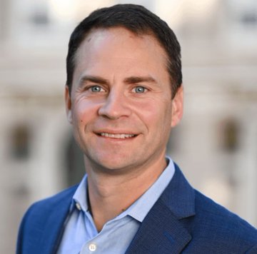 A professional headshot of a smiling middle-aged man with short brown hair and blue eyes, wearing a blue blazer and light blue shirt, photographed against a blurred classical building backdrop.