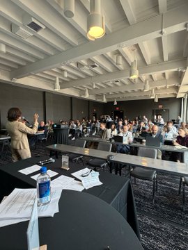 A conference room filled with attendees seated at long tables listening to a speaker, set up for the Humanitarian Payments Council meeting in Washington, D.C., where organizations like Mastercard, Circle, UNDP, and Mercy Corps are gathering to discuss humanitarian financial solutions.
