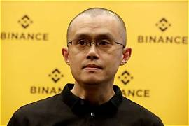 A man with glasses wearing a black jacket stands in front of a yellow backdrop displaying the Binance logo.