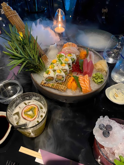 A luxurious sushi platter featuring rolls, sashimi, and garnishes is dramatically presented on a bed of ice with dry ice fog, surrounded by a candle, glasses, and other dishes on a dark table. The "Can I get a GM?" caption pairs perfectly with the extravagant, restaurant-worthy spread that seems worthy of all the engagement it can get.